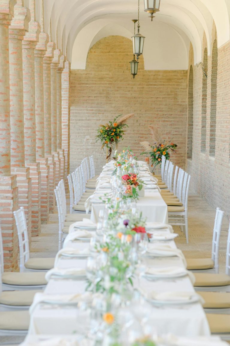 Elegant long table adorned with floral arrangements in a sunlit arched hallway for a wedding or event.